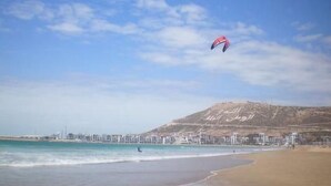 Una playa con cielo despejado, una persona practicando kitesurf y una montaña distante.