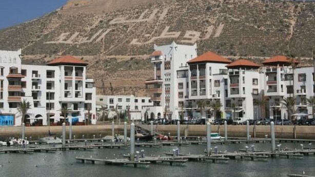 Un puerto deportivo con varias embarcaciones amarradas, rodeado de edificios blancos con tejados rojos y un paisaje montañoso de fondo.