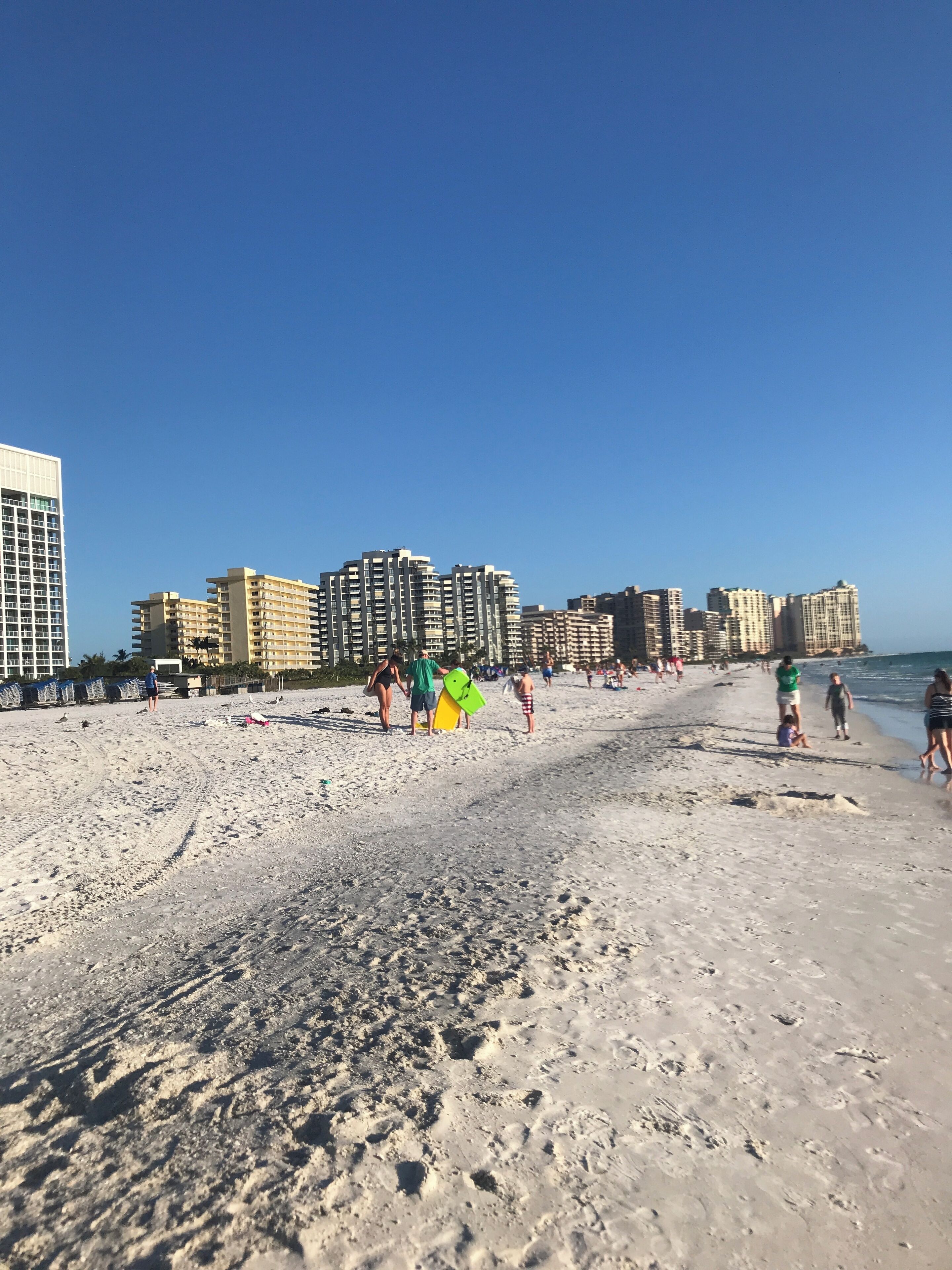 A beach with people walking and standing, and high-rise buildings in the background.