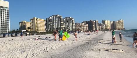 A beach with people walking and standing, and high-rise buildings in the background.