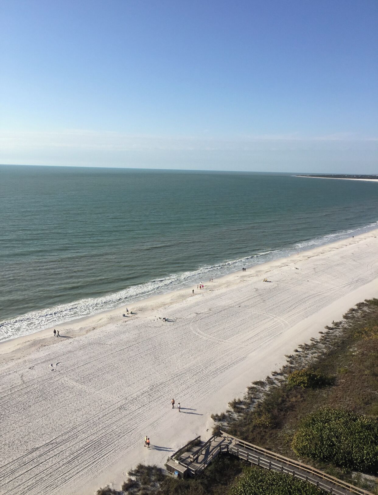 A beach with a long stretch of sand, a few people walking, and a structure in the foreground.