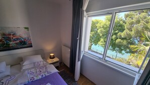 A bedroom with a bed, a nightstand, a lamp, a window with a view of trees and water, and a painting on the wall. - Wohnung 10m vom Strand entfernt by Interhome (Crikvenica)
