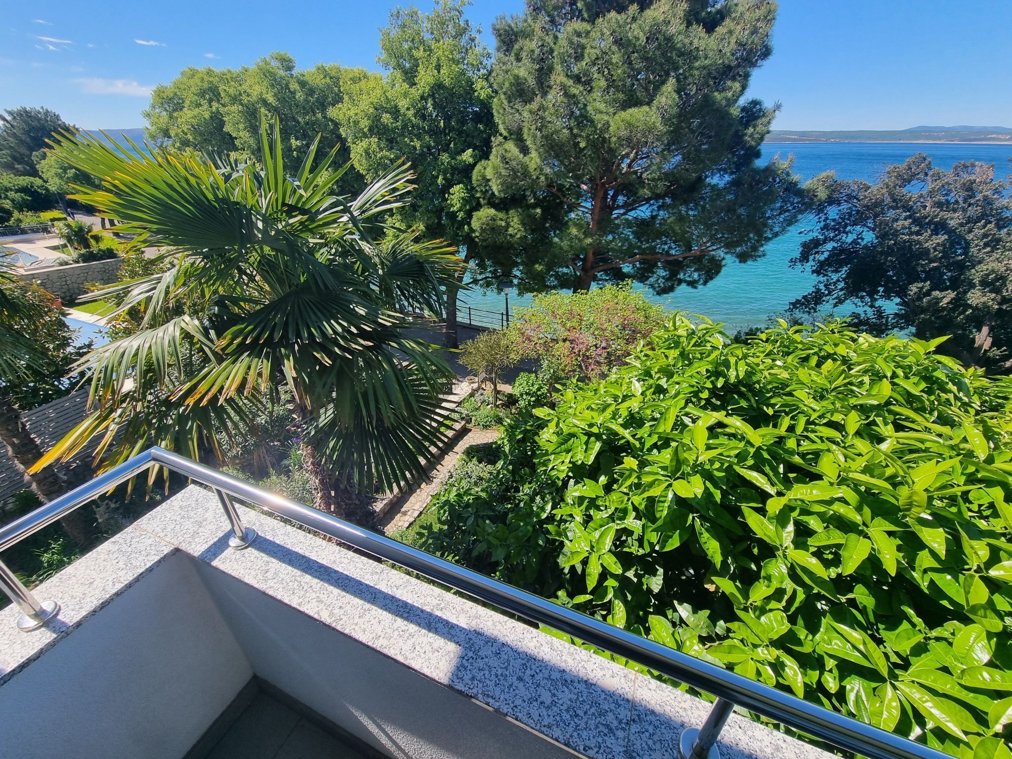 En balkon med udsigt over et kystlandskab, med træer og en klar blå himmel.