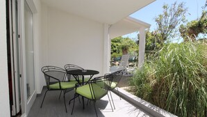 Een overdekt terras met een tafel en stoelen, uitzicht op groen en de ingang van een gebouw.