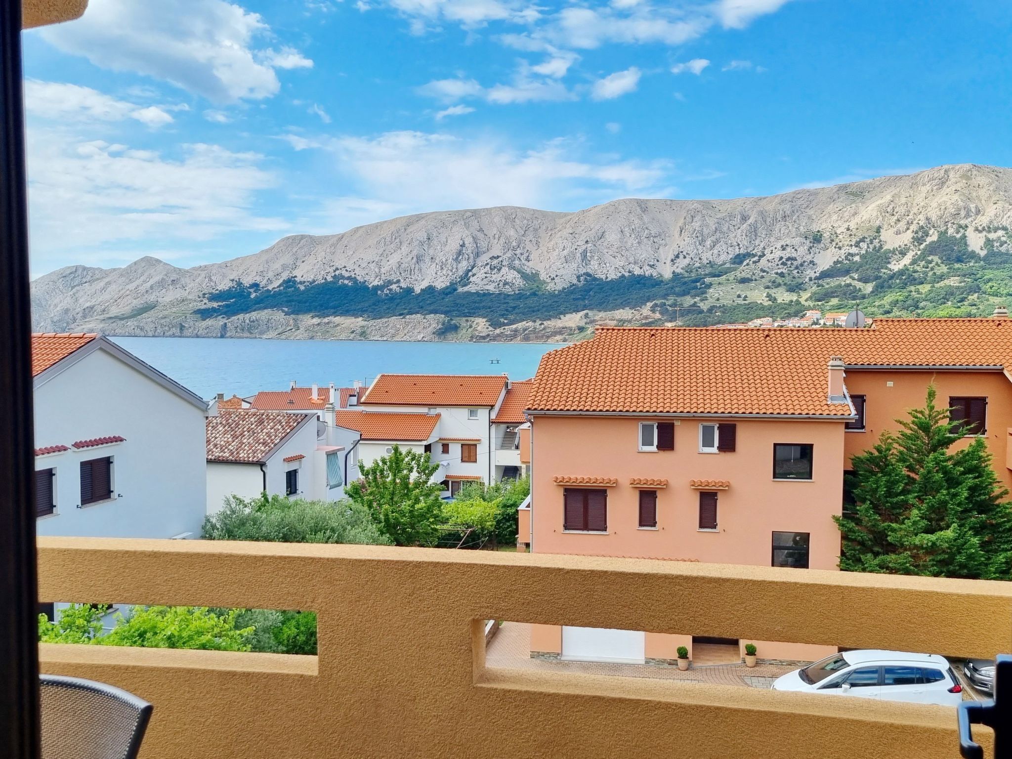 A coastal town with terracotta-roofed buildings, a clear blue sky, and a mountain in the background.