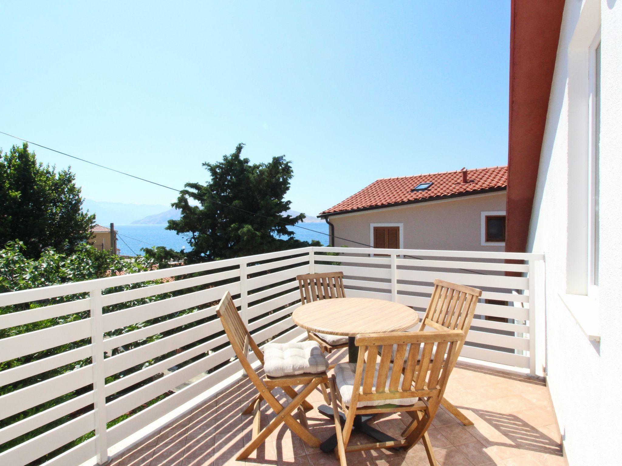 Un balcon avec une table ronde et quatre chaises en bois, donnant sur une zone côtière avec un bâtiment aux toits de tuiles rouges et une vue sur la mer.