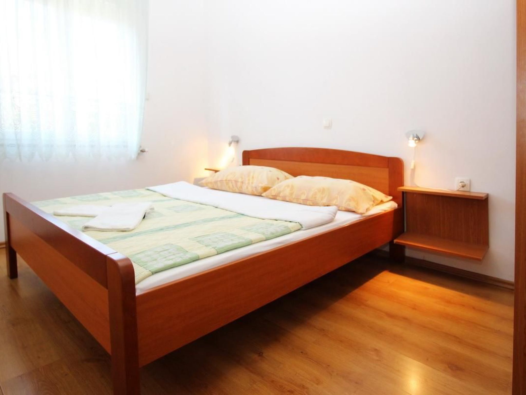 Een eenpersoonskamer met een houten hoofdbord, een nachtkastje met een lamp en een raam met dunne gordijnen.