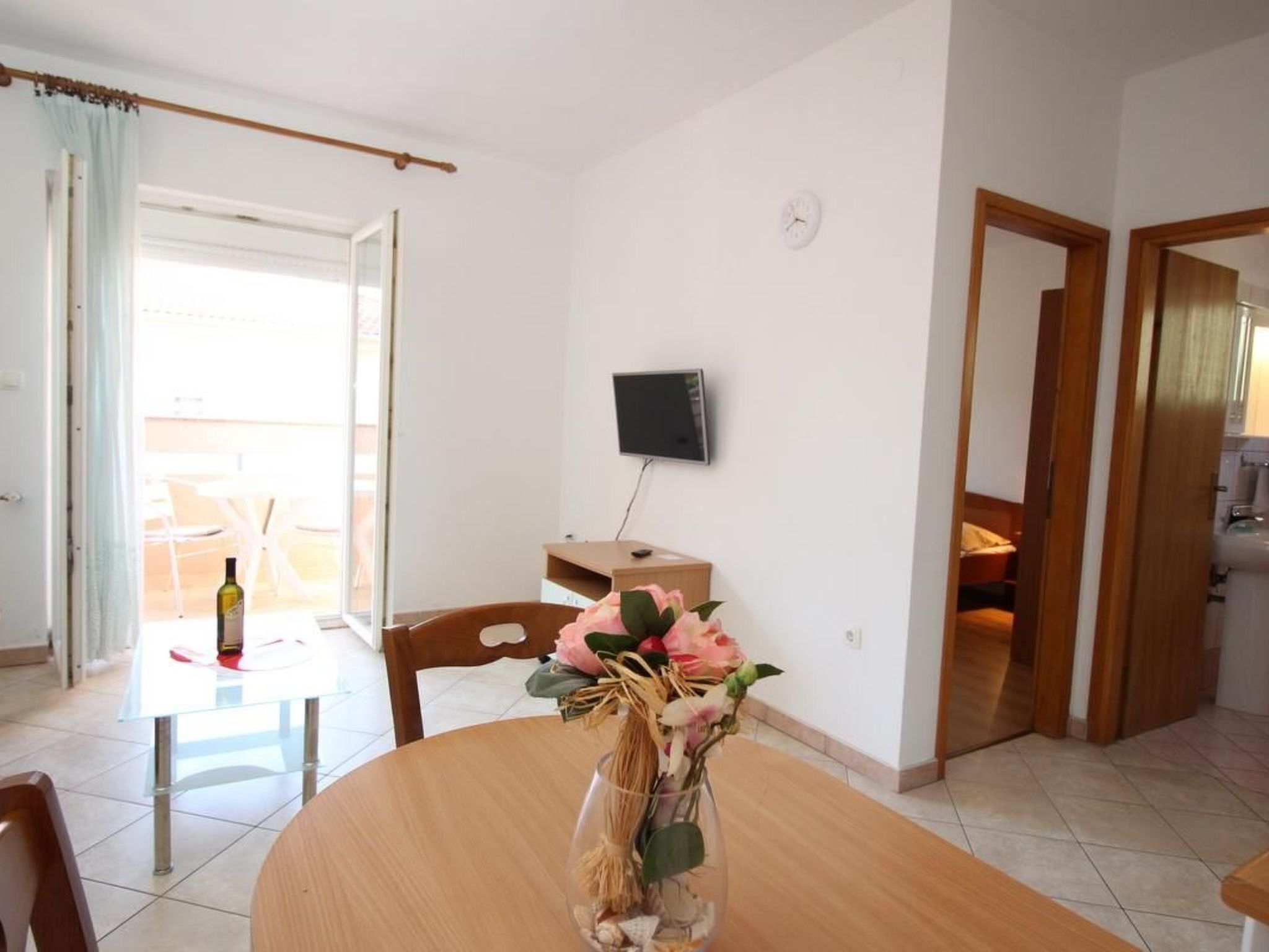 Un coin repas avec une table en bois, des chaises, un vase de fleurs, une bouteille de vin et un téléviseur fixé au mur.