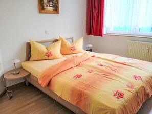Een bed met bloemenbeddels, een bedlampje, een nachtkastje, een raam met gordijnen en een aan de muur bevestigd schilderij.