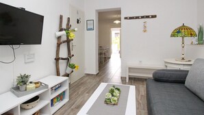 Un salon moderne avec un téléviseur à écran plat, un support à plantes en bois, une table basse grise, un canapé gris et une petite table avec une lampe.