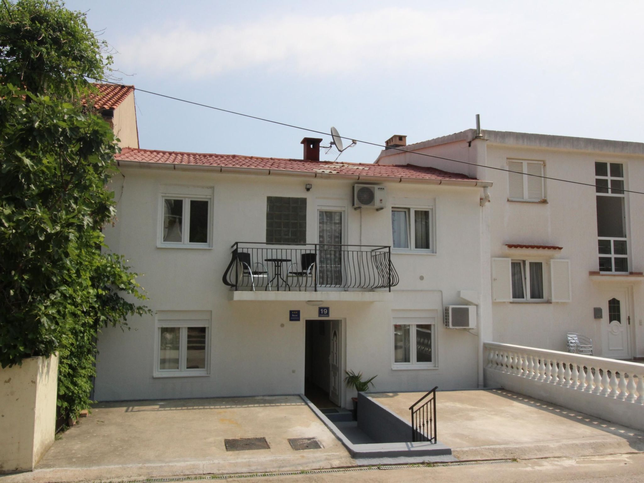 A two-story building with a balcony, a small garden area, and a visible number '19' on the facade.