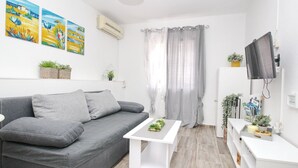 Un salon moderne avec un canapé gris, une table basse blanche et un téléviseur posé sur un meuble. Trois tableaux ornent le mur, une fenêtre est agrémentée de rideaux et une petite plante est posée sur la table basse.
