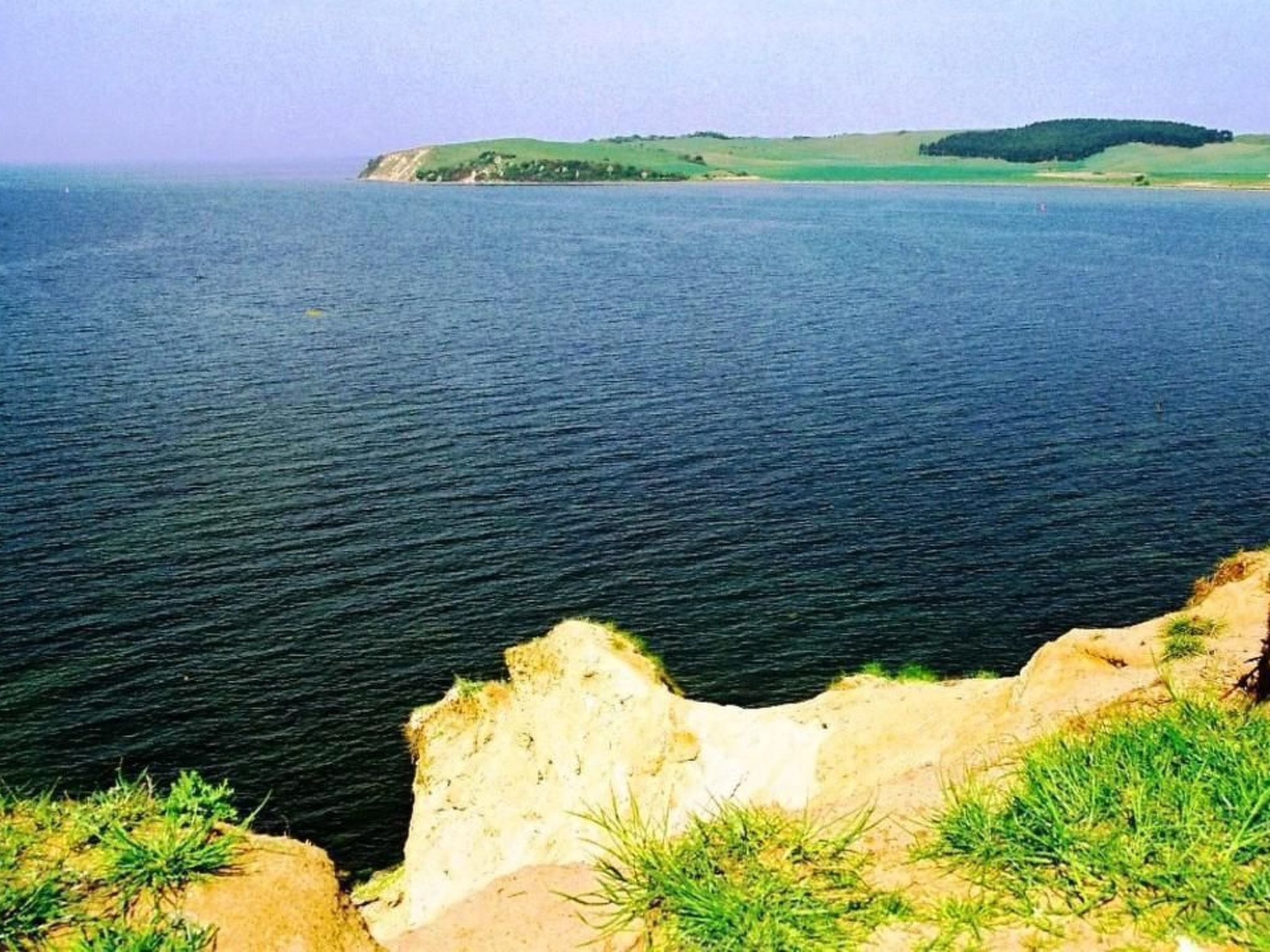 Un paysage côtier avec une falaise, une mer calme et des collines verdoyantes à l’horizon.