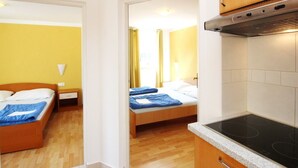 Ett kompakt hotellrum med två sängar, ett litet kök och en väckarklocka på väggen.