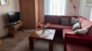 Een woonkamer met een televisie, een houten kast, een rode bank en een tafel met een kantklos.