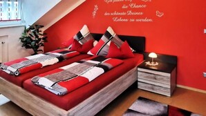 A modern bedroom with a red accent wall, a large bed with multiple pillows, a bedside table with a lamp, and a wall decal with German text. - Fewo im OG mit Balkon by Interhome (Gleißenberg)