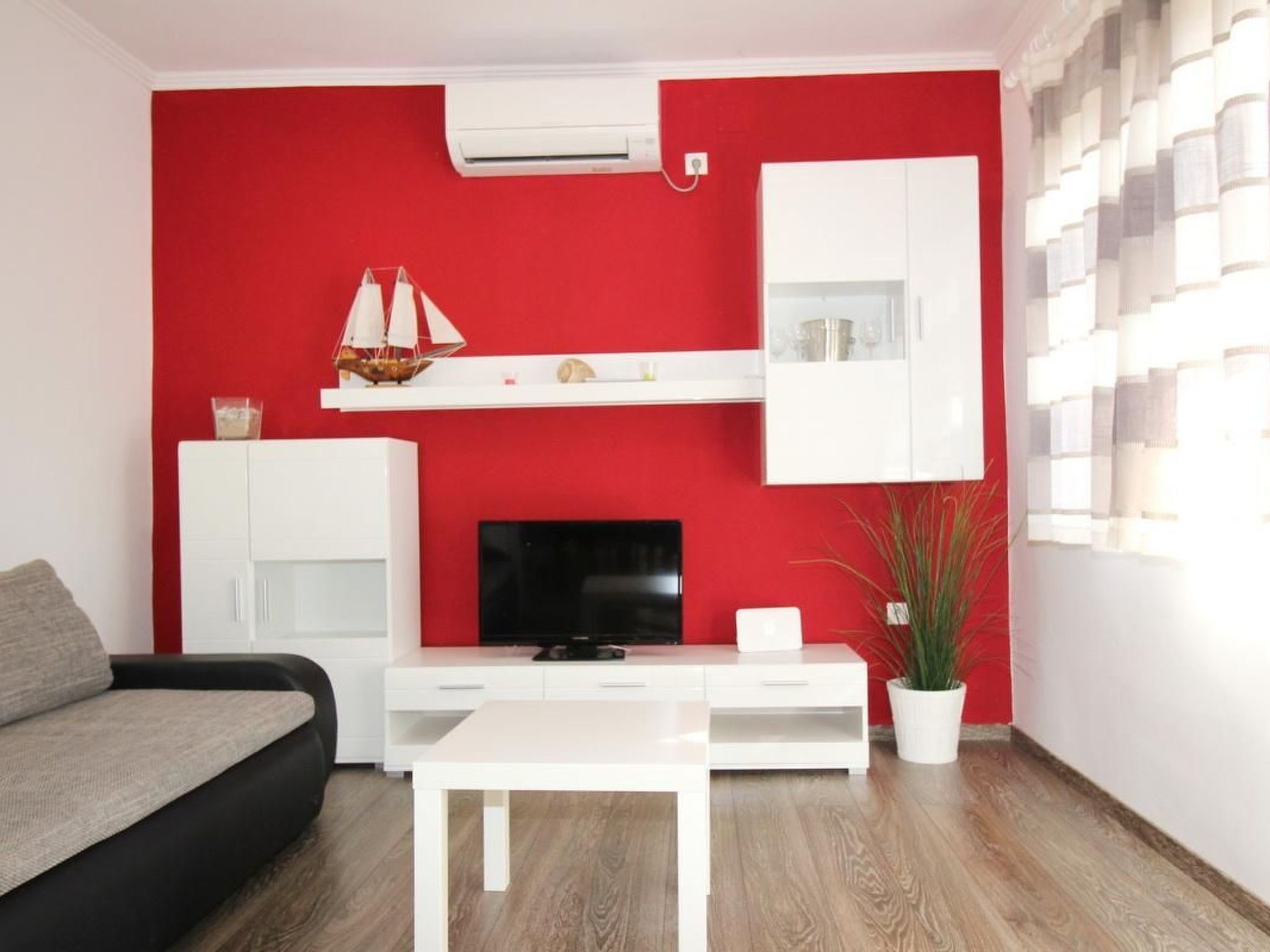 A modern living room with a red accent wall, a flat-screen TV, a white entertainment unit, a grey sofa, and a wooden floor.
