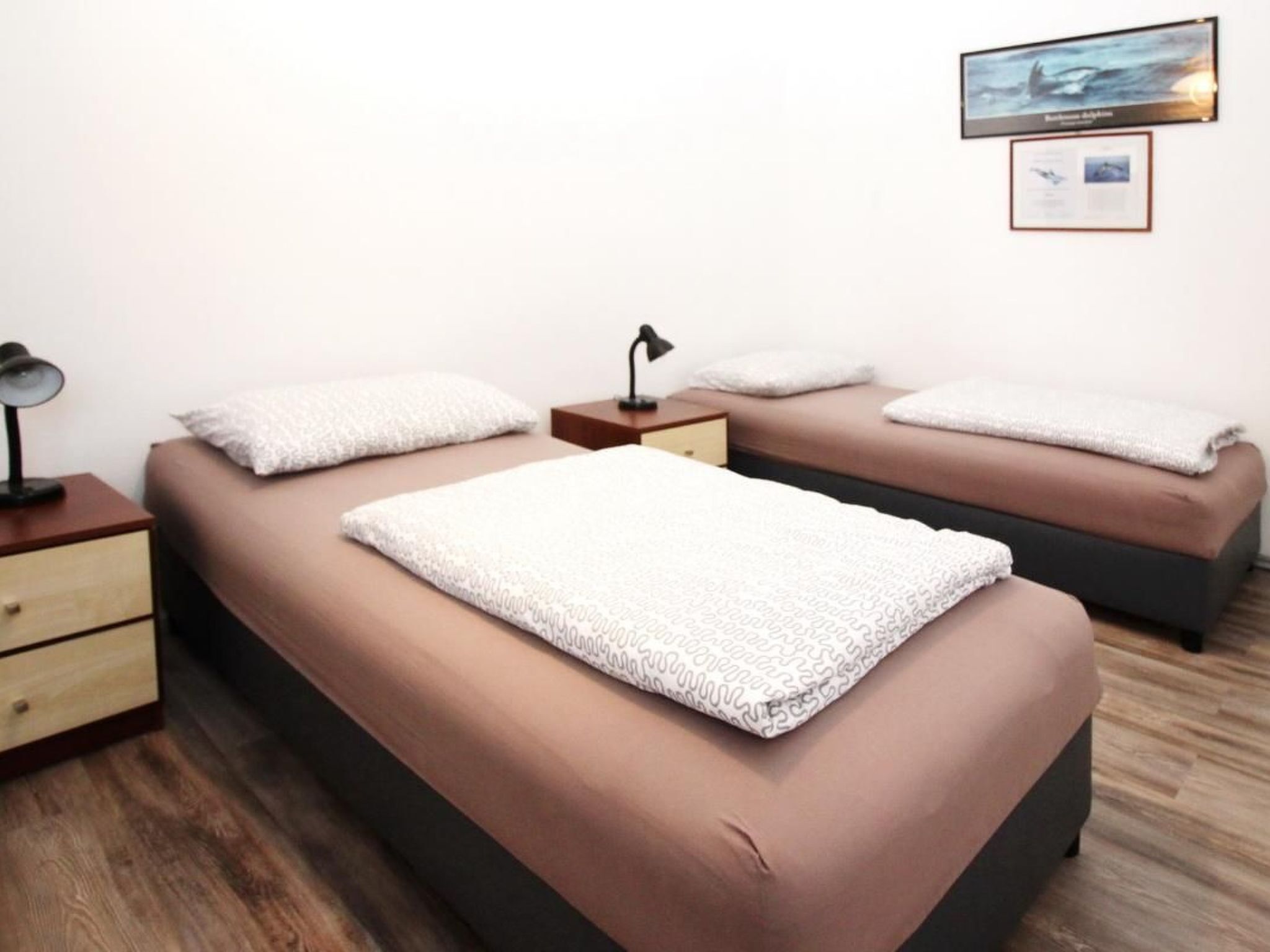 Une chambre avec deux lits, chacun étant équipé d’un oreiller et d’une couverture. Il y a une table de chevet avec une lampe et un tiroir. Un tableau encadré est accroché au mur, au-dessus des lits.