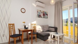 A modern living room with a sofa, two wooden chairs, a small table, and a dining table. There is a clock on the wall, an air conditioning unit, and a painting above the sofa. - Appartement in Baška mit Eigenem Balkon by Interhome (Krk/Baška)