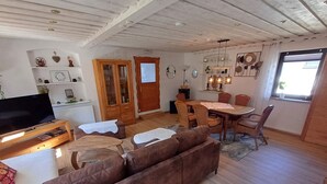 Una sala de estar acogedora con un sofá marrón, una mesa de centro de madera y un mueble para televisión. Hay un comedor con una mesa y sillas de madera, y una zona de cocina con armarios y un ventanal.