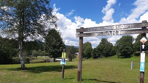 Un cartel de madera con la inscripción 'DREIKONIGSLOIPE START', rodeado de un área cubierta de césped, árboles y un cielo despejado.