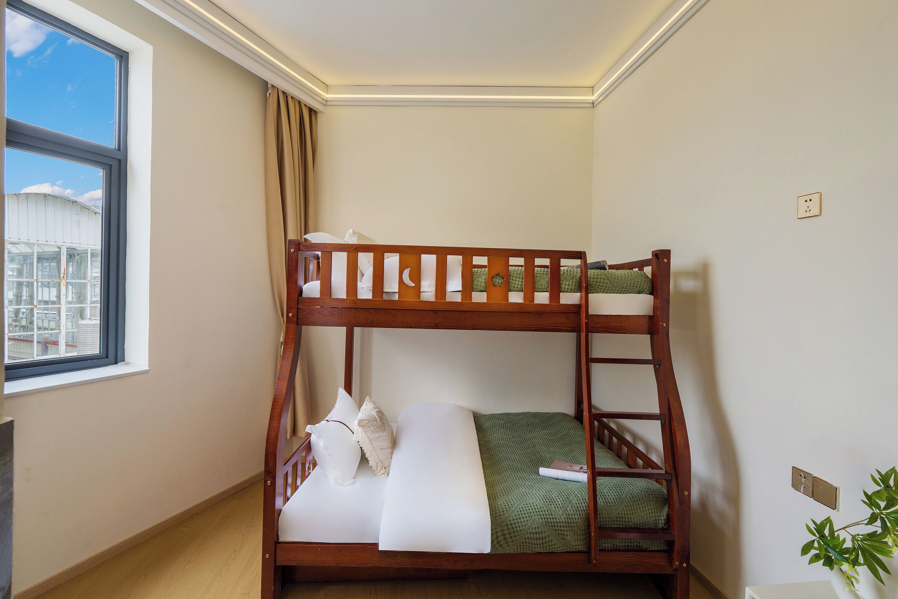 Ein Etagenbett mit weißer Bettwäsche und einer grünen Decke, ein Fenster mit Blick auf Gebäude und eine Pflanze auf der rechten Seite.