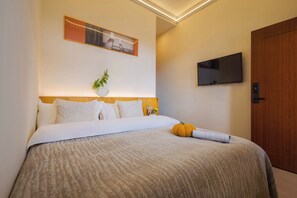 Uma cama bem arrumada com manta texturizada, uma TV fixada na parede, uma porta de madeira e um quadro emoldurado acima da cama.