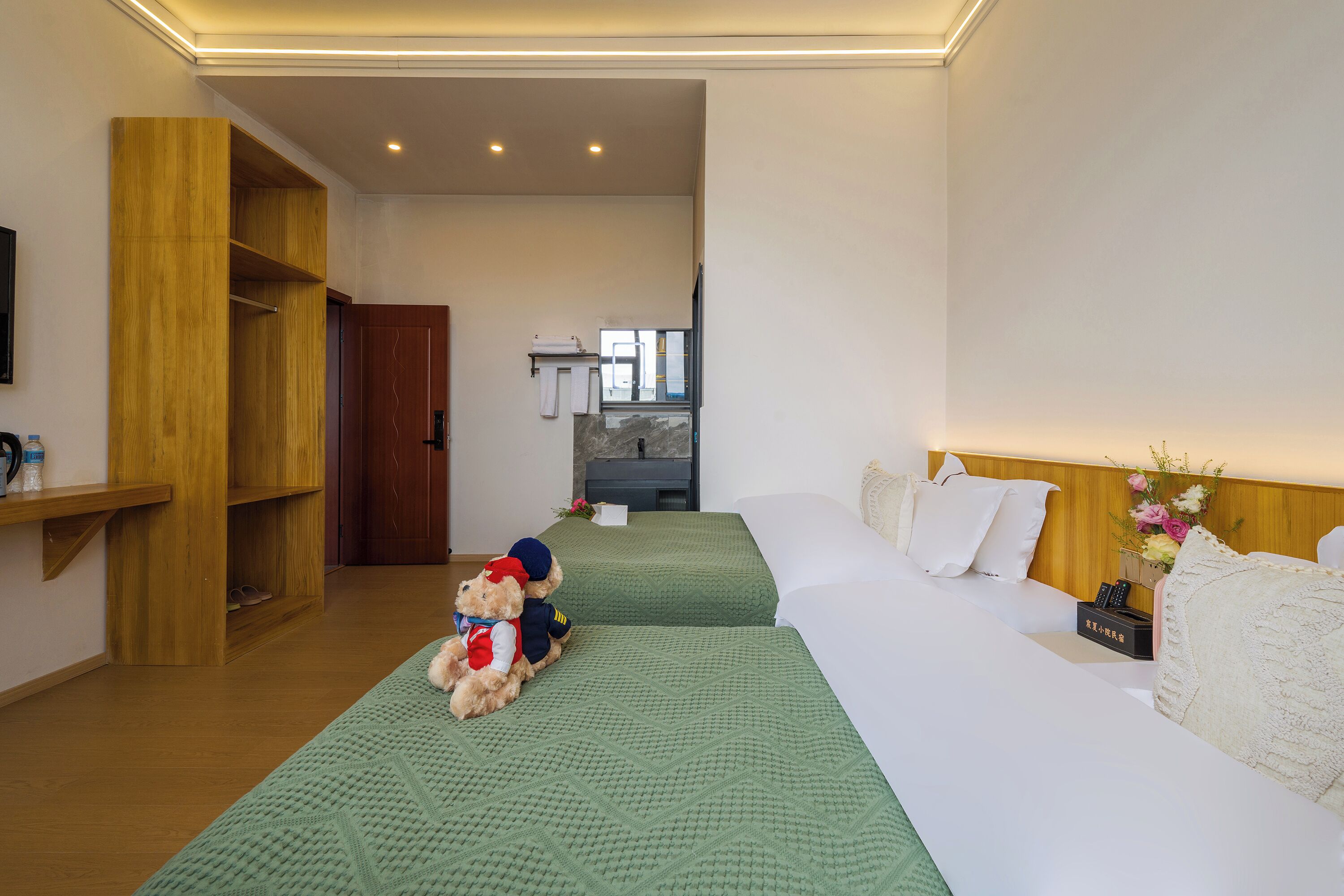 A hotel room with two beds, a wooden shelf, a teddy bear, and a small table with a vase of flowers.