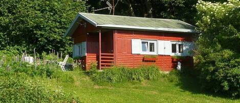 A red wooden cabin with a green roof, surrounded by lush greenery and trees.