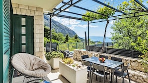 Un patio con una mesa y sillas, un sillón acolchado, una mesita con tazas y vistas al mar y a las montañas.