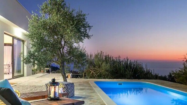 A modern outdoor pool area with a lounge chair and a table, surrounded by a tree and a view of the sea at sunset. - Stylische Villa -Tres Chic- in Makarska by Interhome (Makarska)