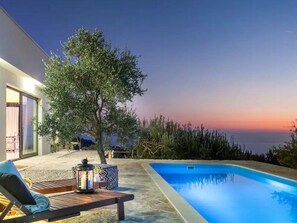 Un área de piscina moderna al aire libre con una silla de descanso y una mesa, rodeada por un árbol y una vista del mar al atardecer.