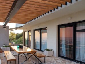 Un patio exterior con mesa y bancos de madera, ventanas amplias y plantas en macetas.
