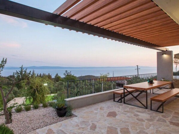 Una terraza con suelo de piedra, mesa y bancos de madera, plantas en macetas y vista al mar.