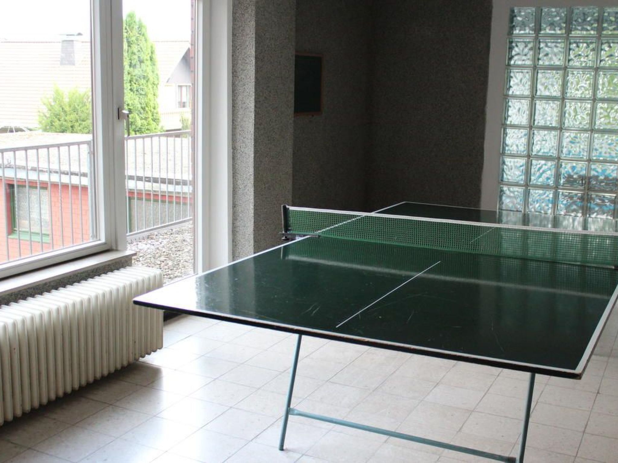 A ping pong table in a room with a large window, a radiator, and a view of a building outside.
