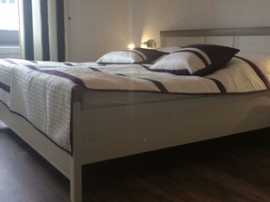 Uma cama com colcha acolchoada e almofadas, uma mesa de cabeceira com abajur e uma janela com persianas.
