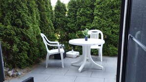 En udendørs patio med et rundt bord og to hvide stole, omgivet af frodigt grønt.