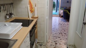 Et køkken med vask, komfur og diverse køkkenredskaber. En dør fører ind til en opholdsstue med sofa og udsigt udad.