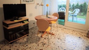 En stue med et fladskærms-tv på et træstel, et rundt bord med stole, en vase med blomster og et billede på væggen.