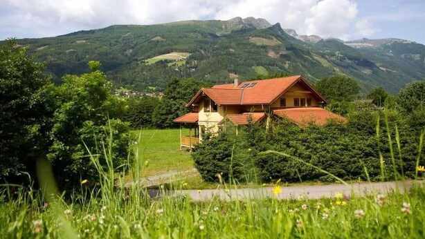 Et hus med rødt tag omgivet af frodig vegetation og bjerge i baggrunden.