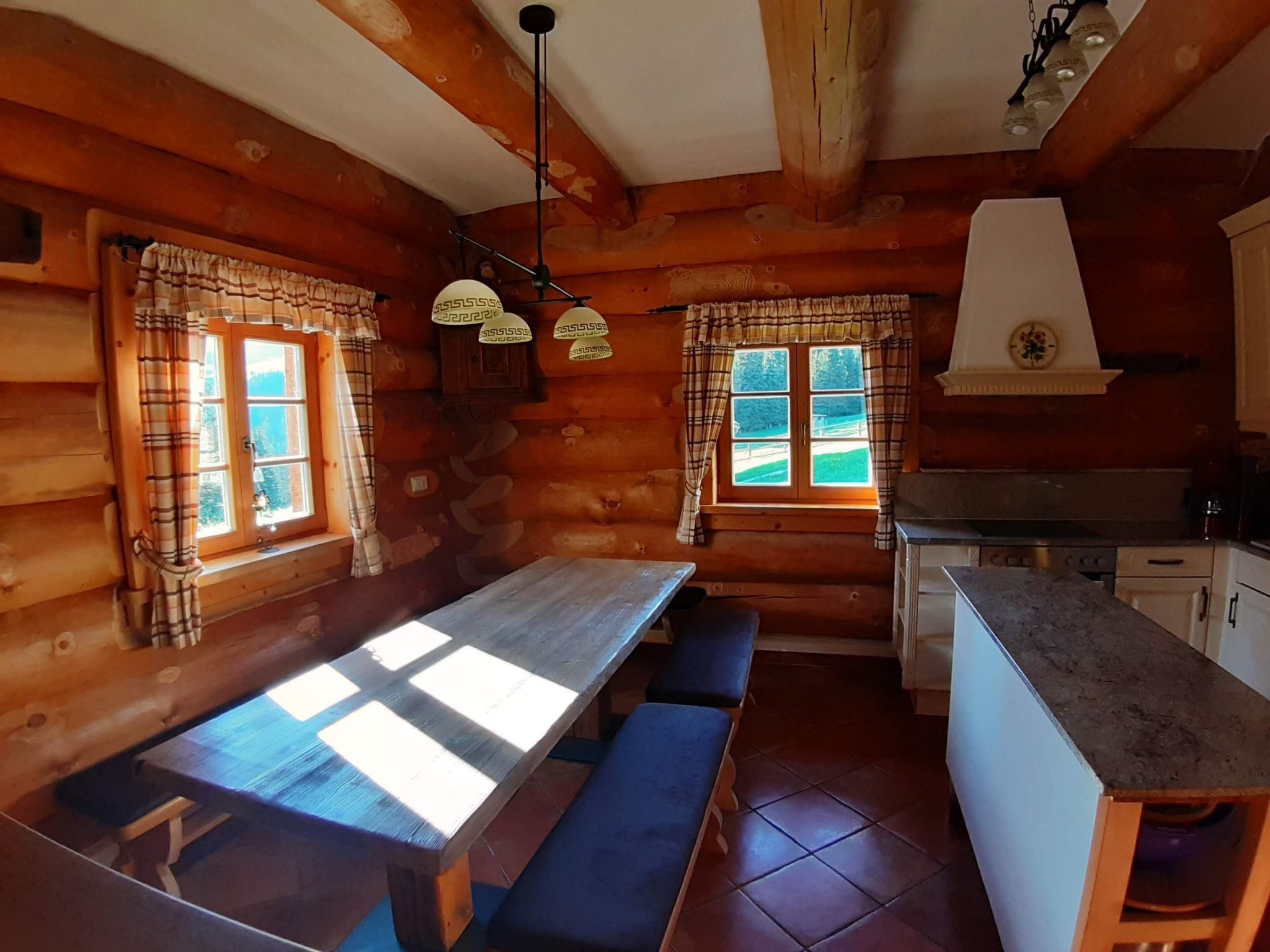 Ein Blockhaus-Interieur mit einem Holzespeisetisch und Bänken, einer Küchenzeile und großen Fenstern mit Karottenvorhängen.