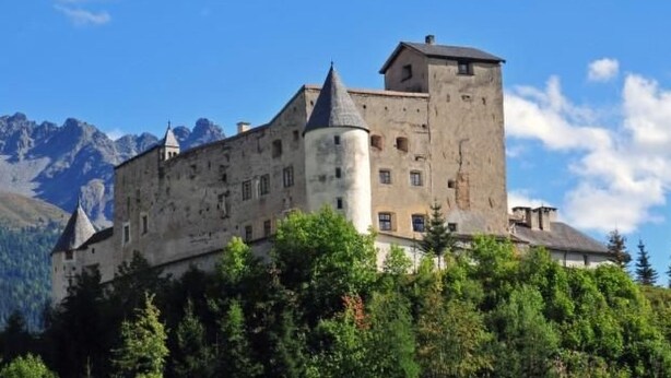 Eine historische Burg mit einem markanten Turm, umgeben von üppiger Vegetation und Bergen im Hintergrund.