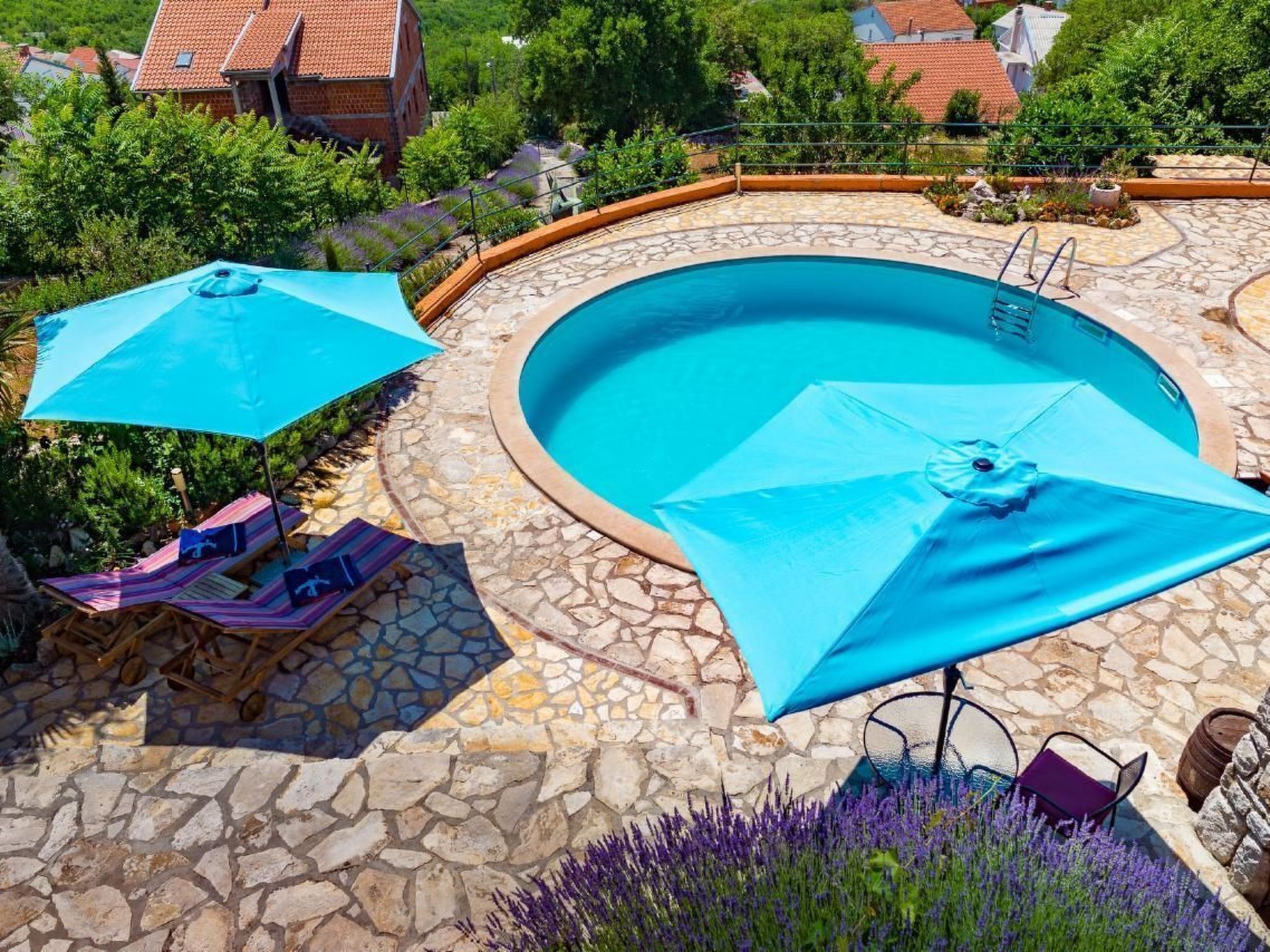 A circular pool with stone tiles, surrounded by a stone patio, two blue umbrellas, and lounge chairs.