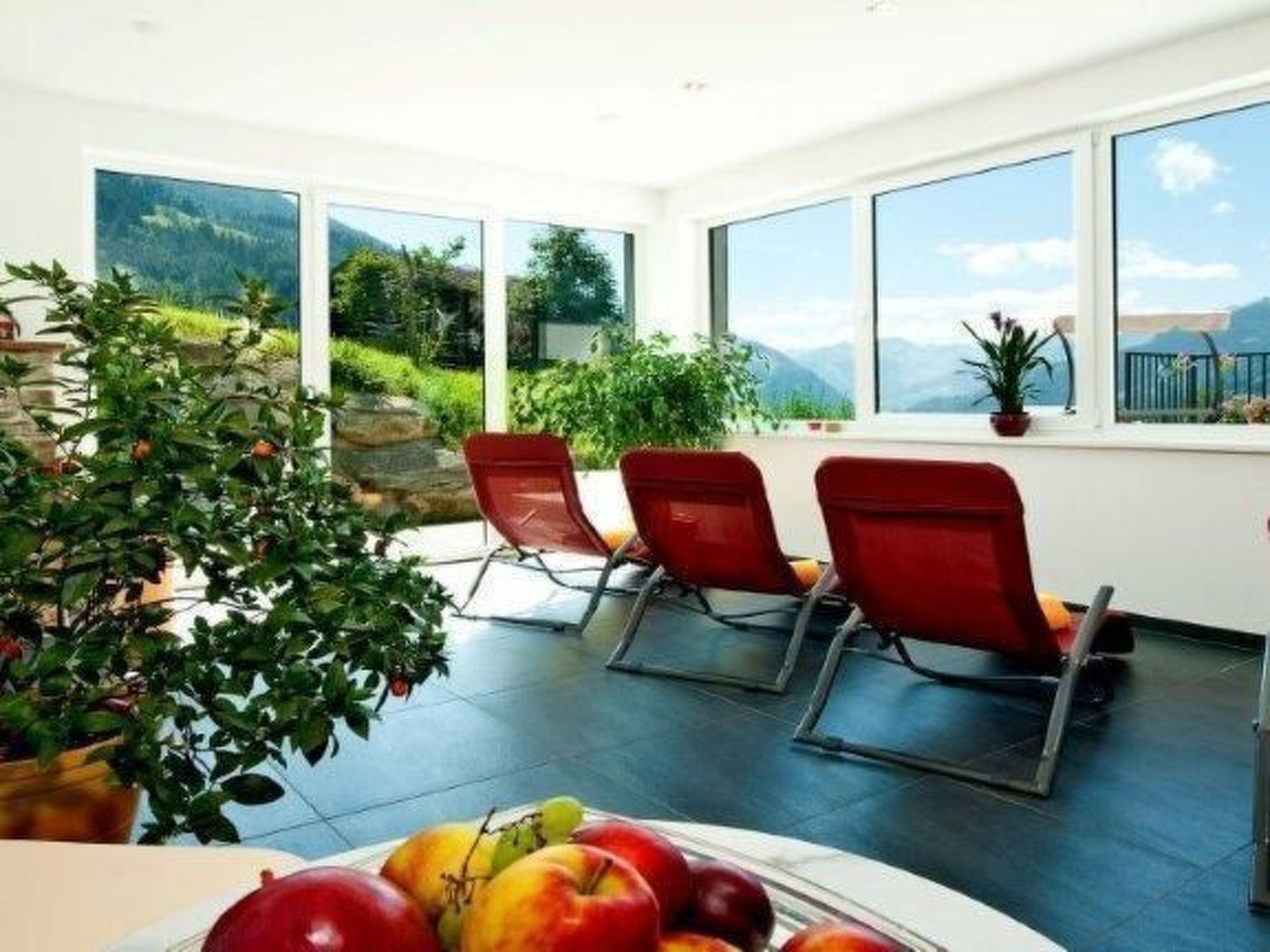 A room with large windows, red chairs, a table with fruit, and a view of mountains.