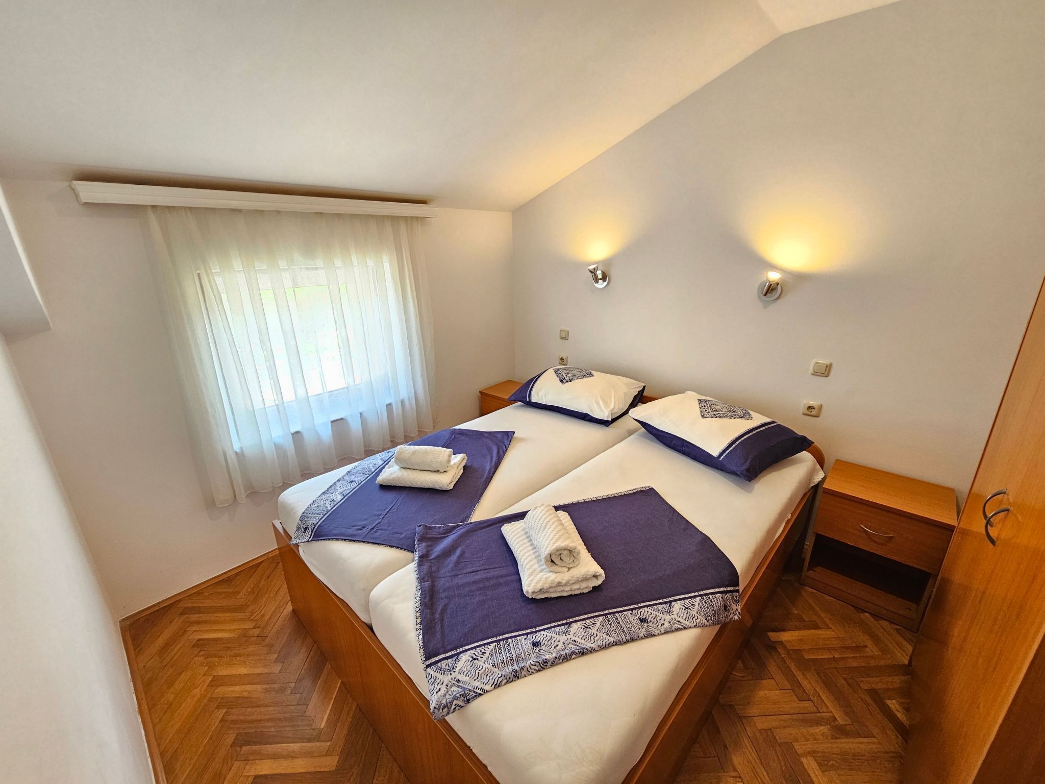 Un dormitorio con dos camas, piso de madera, ventana con cortinas e iluminación en la pared.