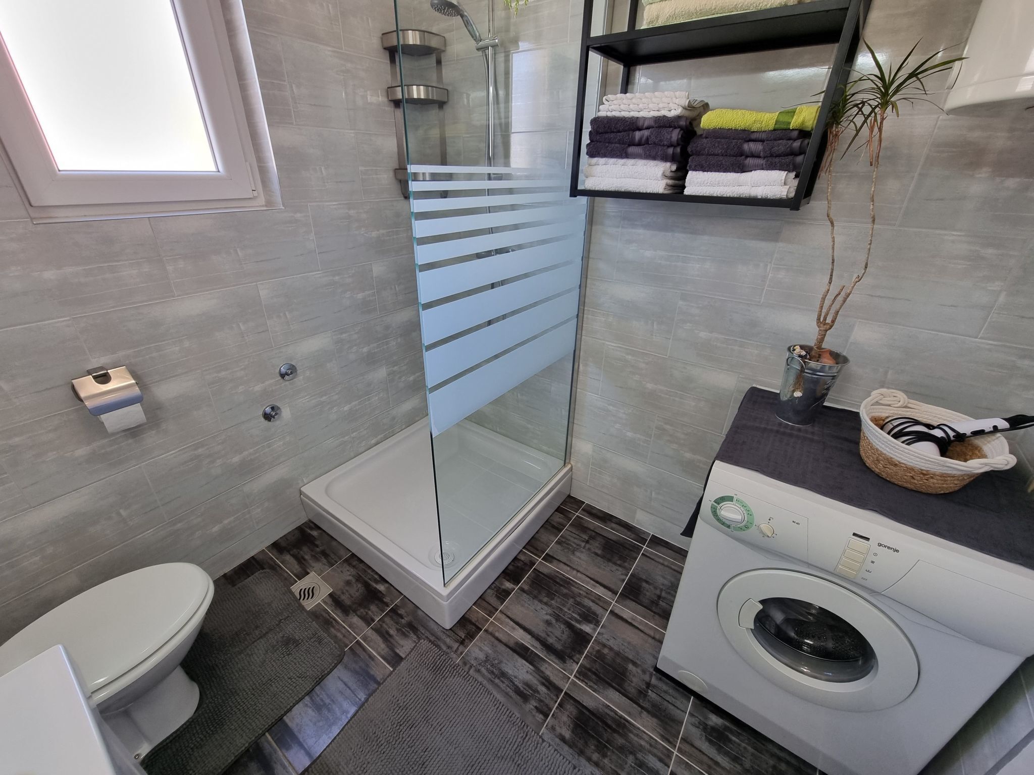 A modern bathroom with a glass shower enclosure, a toilet, a washing machine, and a shelf with folded towels.