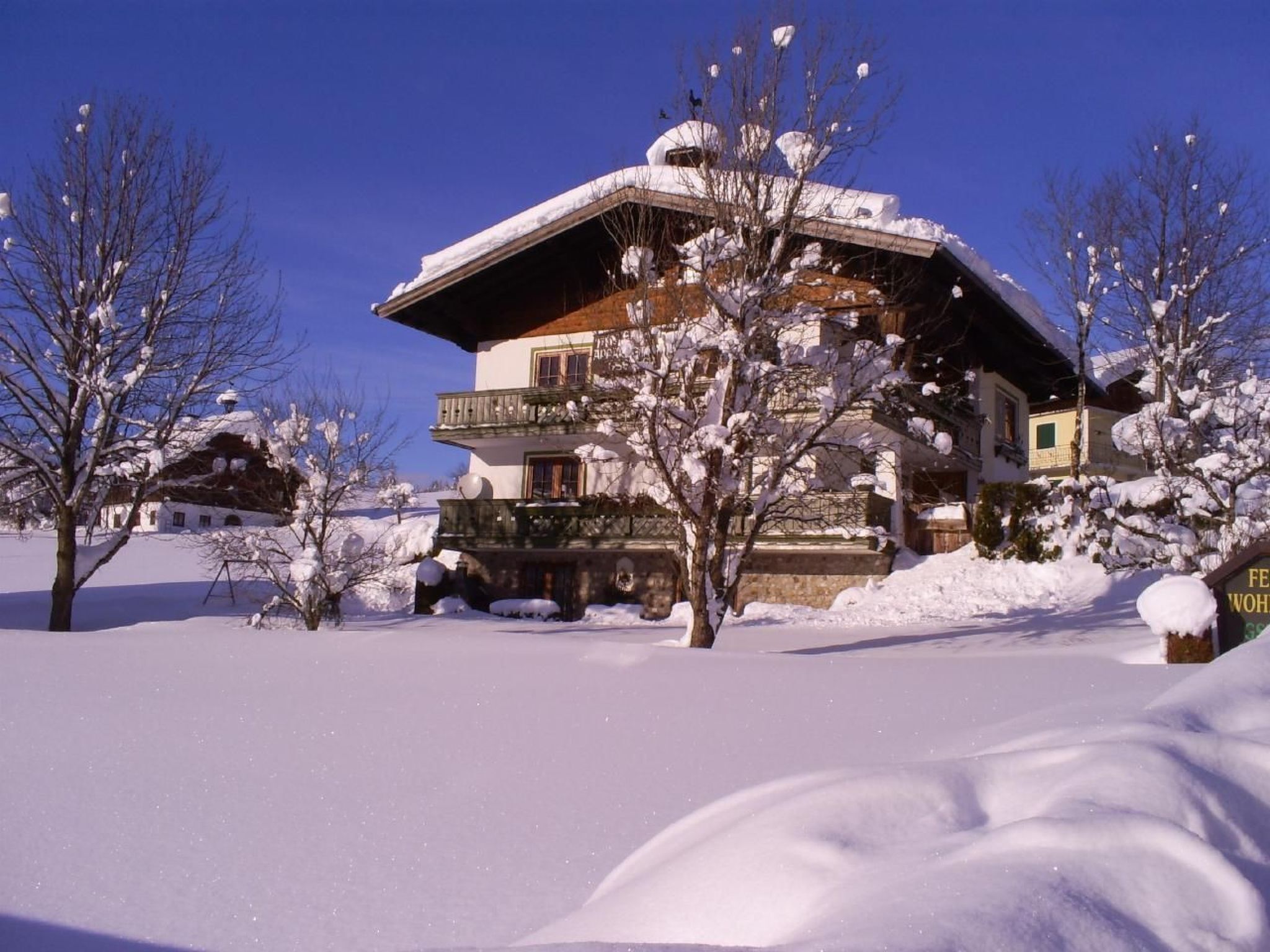 Een chalet met twee verdiepingen, met een met sneeuw bedekte sneeuwdak en balkon, omringd door besneeuwde bomen.
