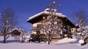 Een chalet met twee verdiepingen, met een met sneeuw bedekte sneeuwdak en balkon, omringd door besneeuwde bomen.