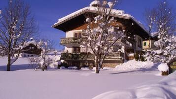 Een chalet met twee verdiepingen, met een met sneeuw bedekte sneeuwdak en balkon, omringd door besneeuwde bomen.