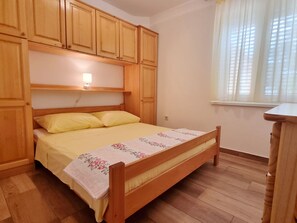 A bedroom with a wooden bed, yellow bedding, a floral bedspread, wooden cabinets, a bedside table, and a window with curtains. - Barbalić by Interhome (Krk/Baška)